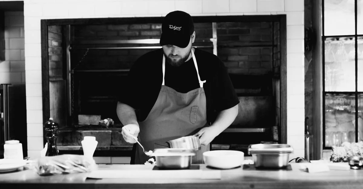 Un chef portant un tablier prépare un repas dans une cuisine rustique. Photographie en noir et blanc.