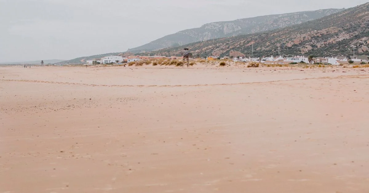 Plage de sable paisible à Cádiz, Espagne, avec des collines lointaines et un village.
