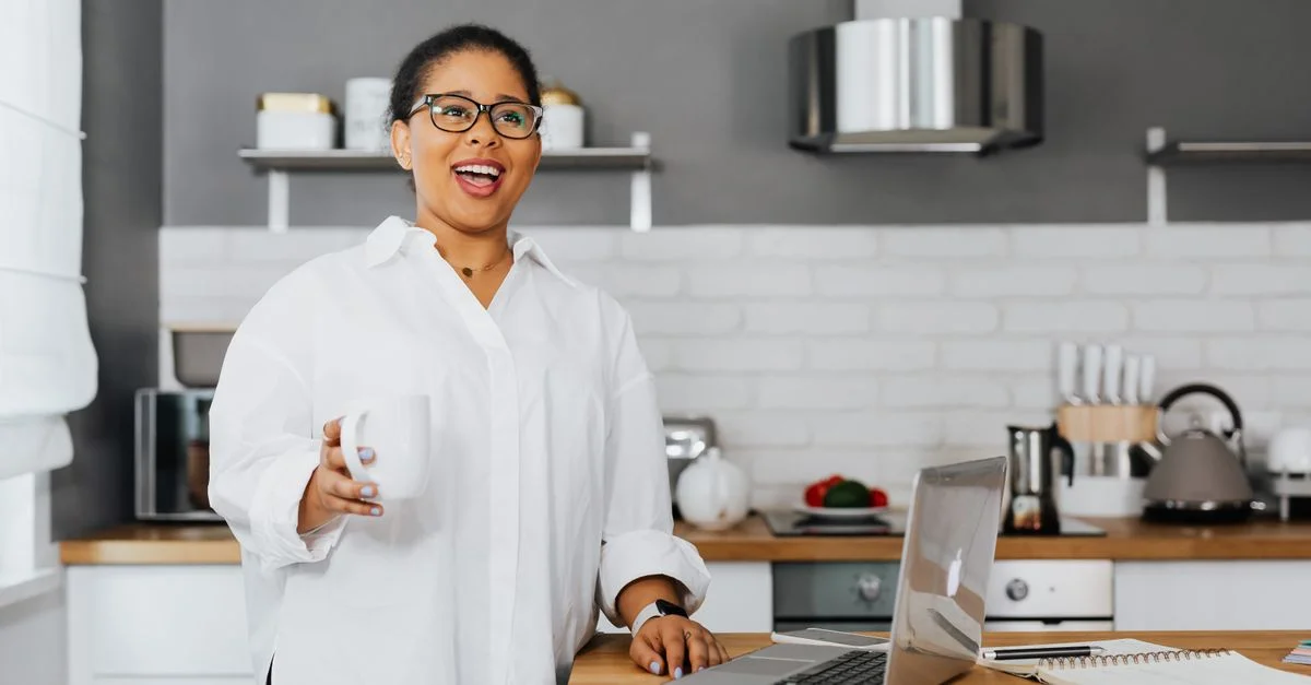 Femme confiante portant des lunettes tenant une tasse, souriante tout en se tenant dans une cuisine moderne avec un ordinateur portable.