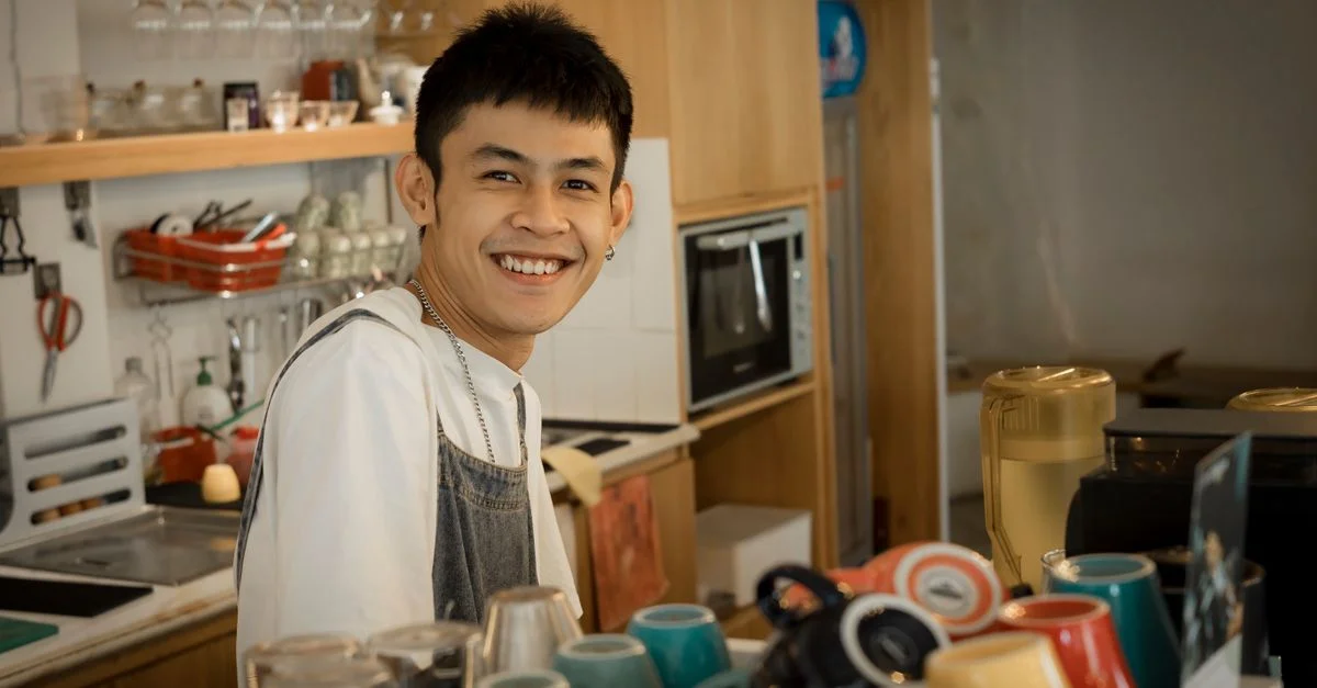 Barista joyeux souriant derrière le comptoir dans un café chaleureux avec des tasses colorées.